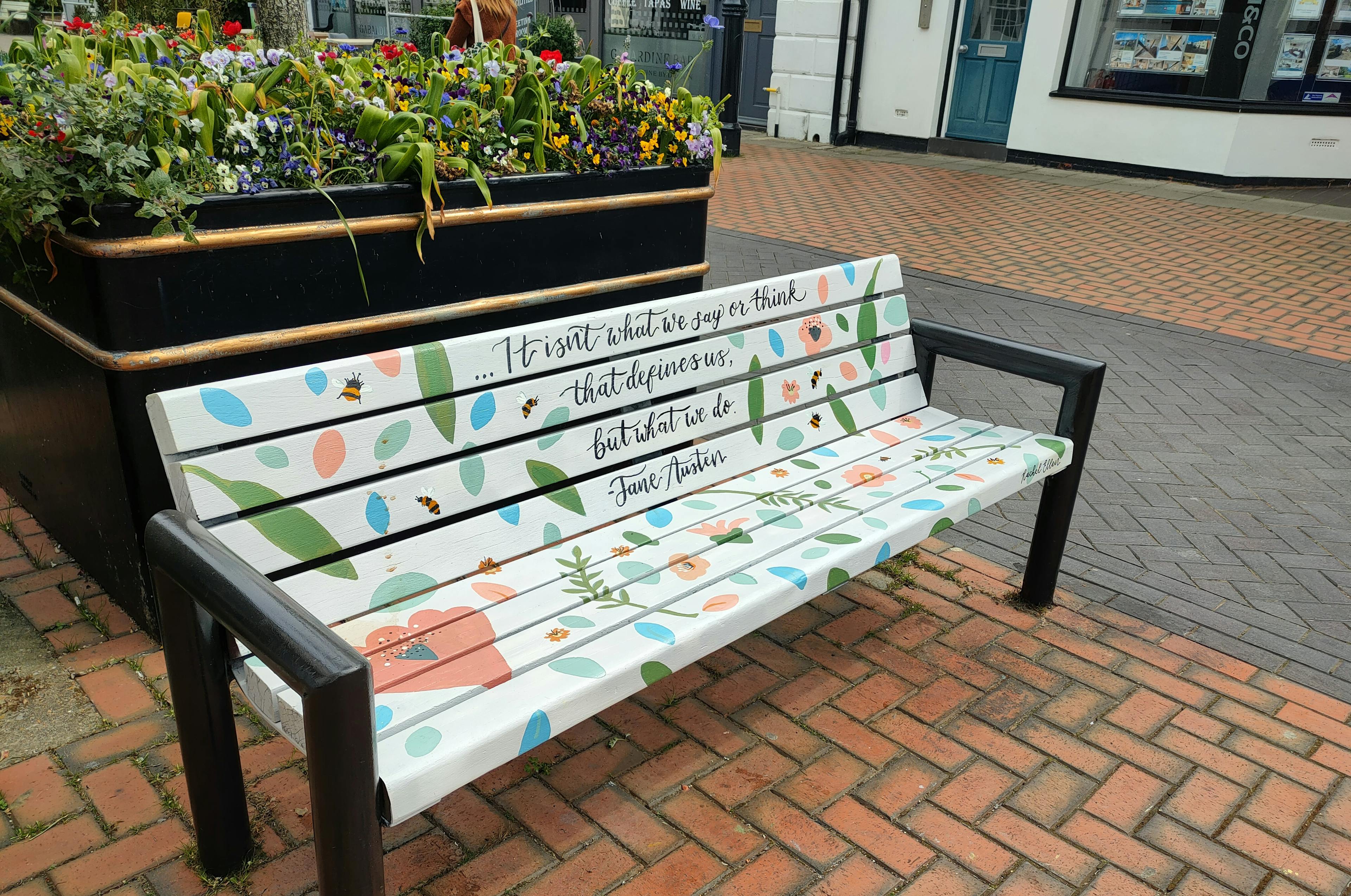 A bench with black arms is painted white with a quote by Jane Austin. It is decorated with leaves and flowers.