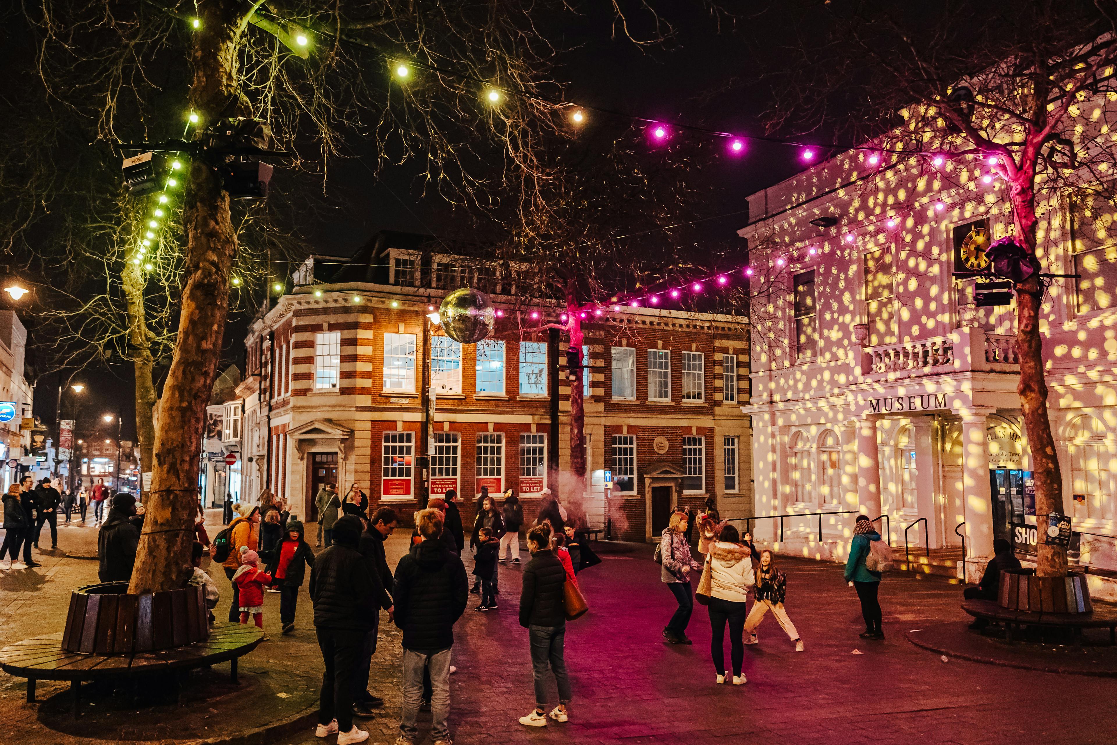 Image shows a crowd gathered outside in a square. There are white light projections on a building, festoon lights in yellow and pink with a glitter disco ball in the middle. The crowd watch the display.